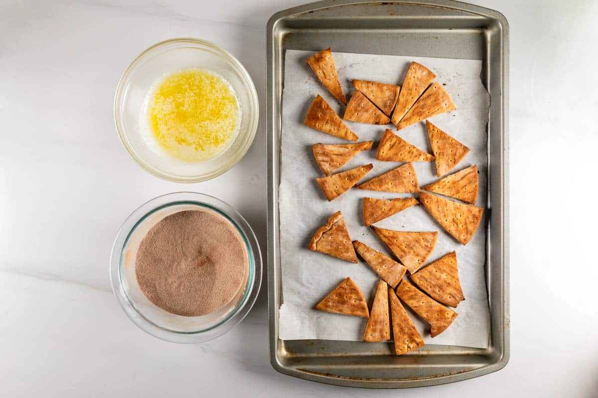 A baking tray with triangular tortilla chips on parchment paper is placed next to a bowl of melted butter and a bowl of cinnamon sugar.