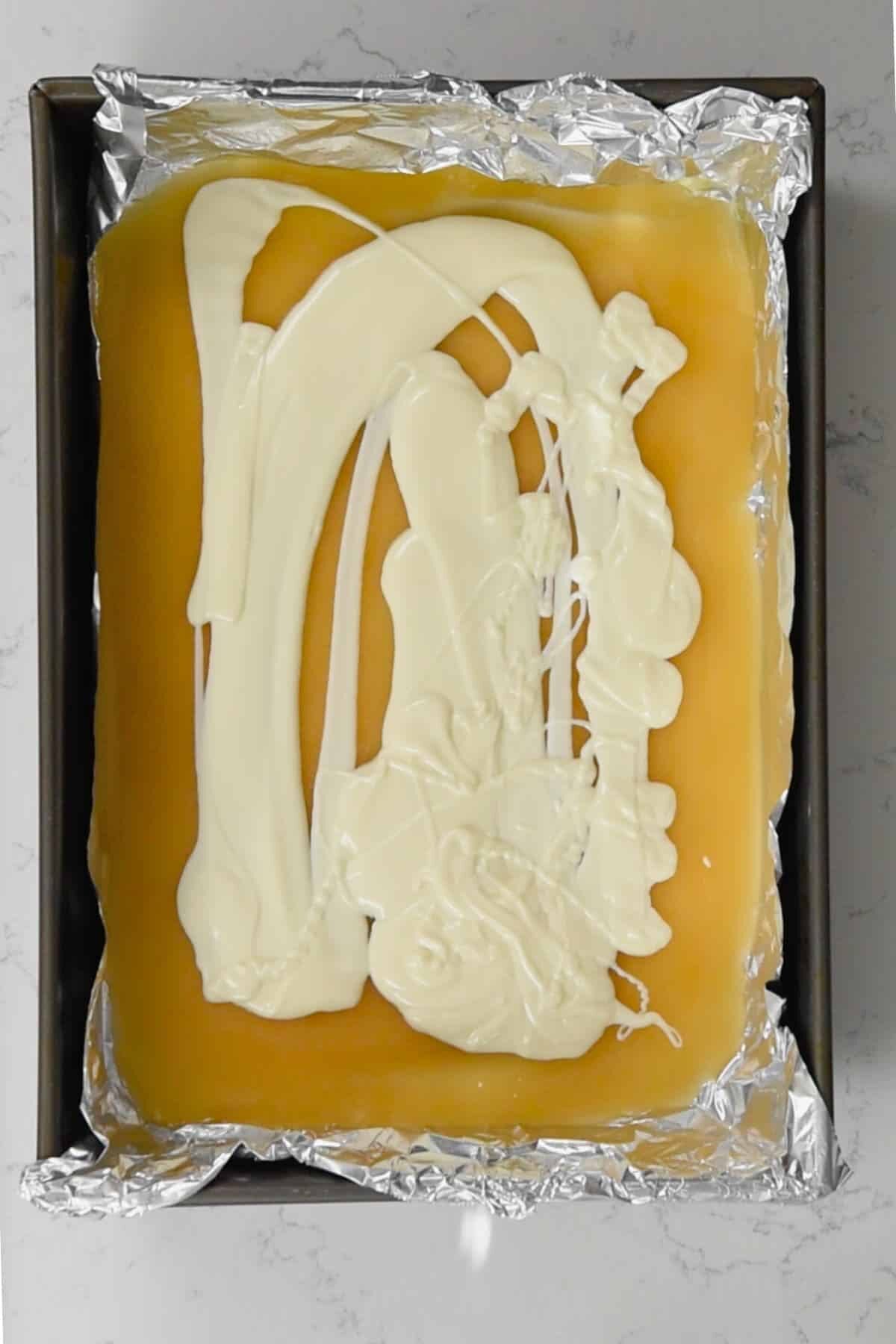 A baking pan lined with aluminum foil contains a smooth layer of churro toffee, topped with a swirl of white chocolate. The pan is set on a light-colored marble countertop.