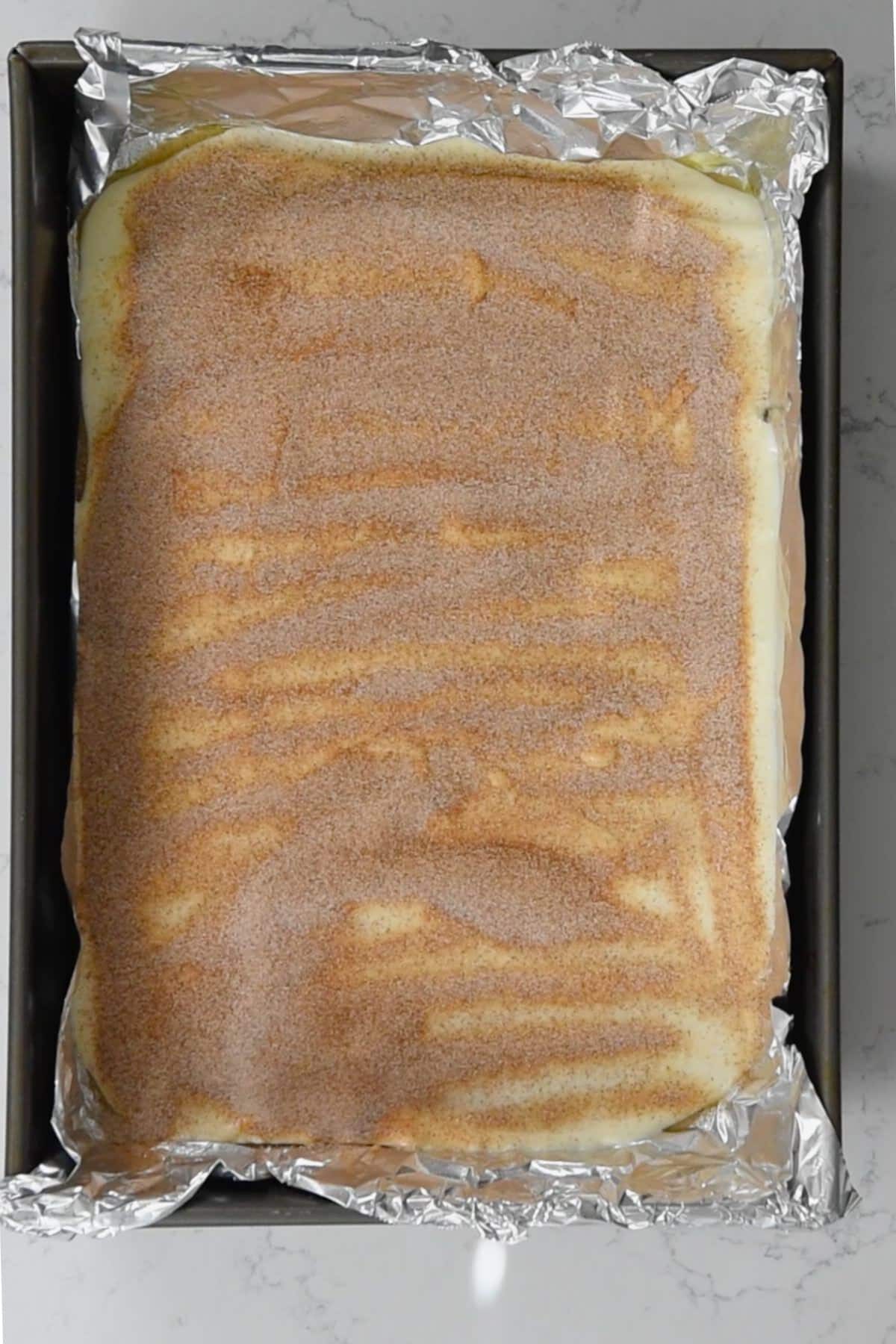 A rectangular baking tray filled with churro toffee is topped with a layer of cinnamon sugar. The tray is lined with aluminum foil, and the dessert boasts a smooth, even top adorned with visible swirls of cinnamon. Its set on a marble countertop.