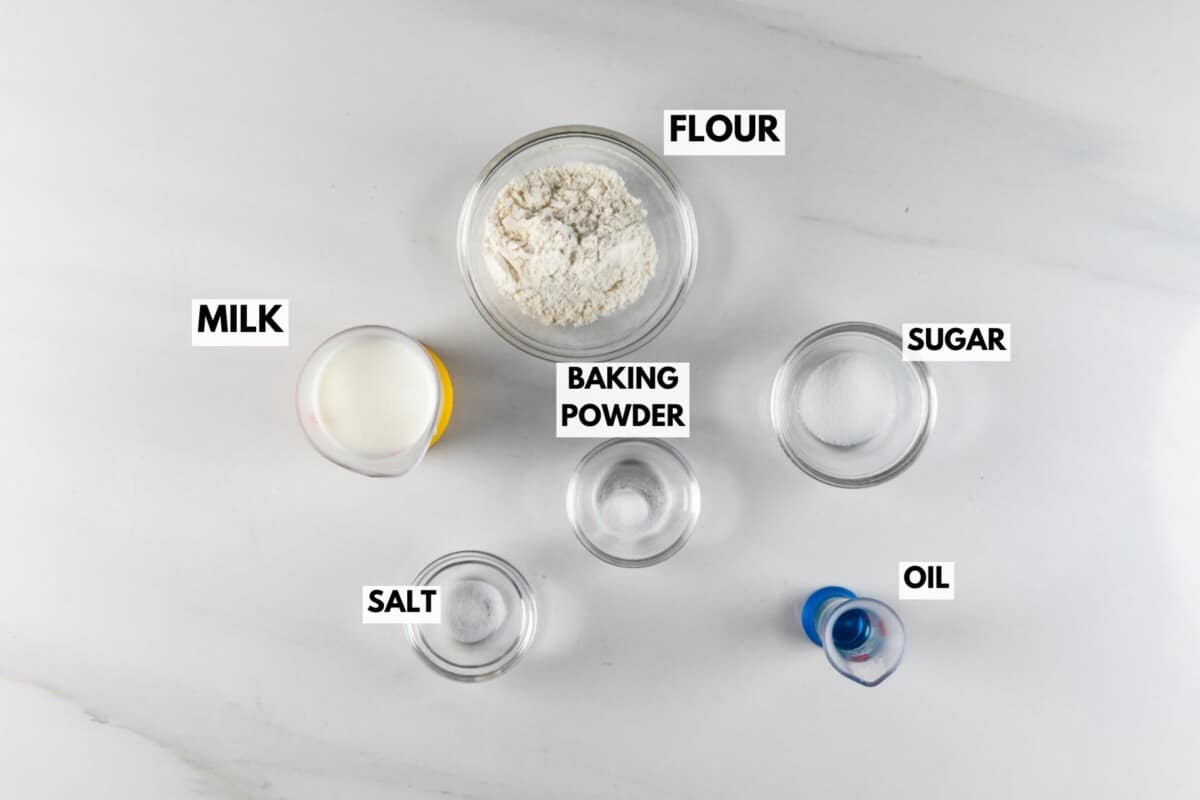 Top-down view of labeled ingredients in small bowls on a white surface: flour, sugar, milk, salt, oil, and baking powder. Each item is placed separately and clearly marked.