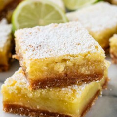 Two lemon bars stacked on a marble surface, dusted with powdered sugar. A slice of lime is visible in the background, adding a hint of green to the scene. The bars have a golden brown crust and a creamy yellow filling.