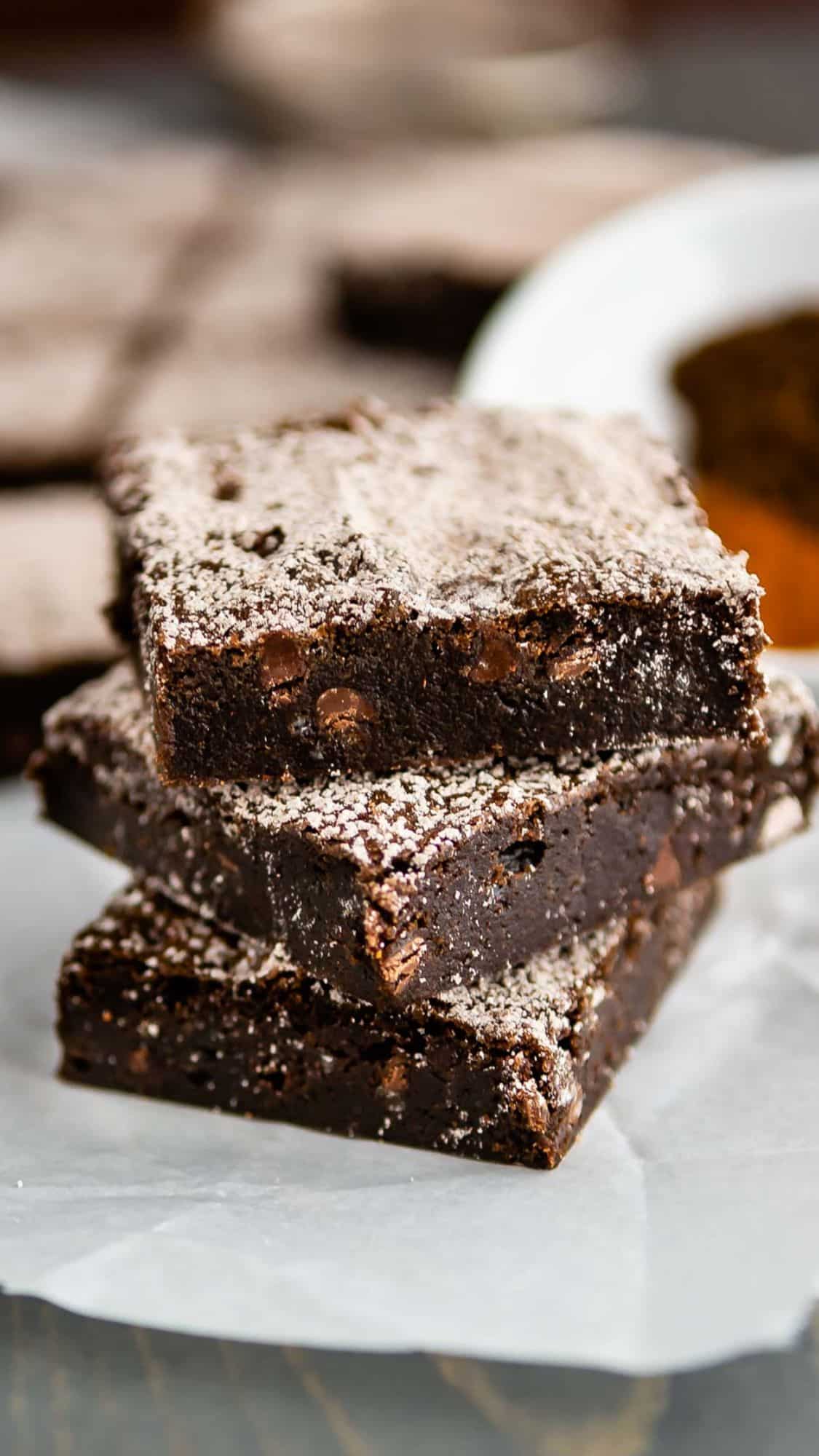 A stack of three chocolate brownies dusted with powdered sugar sits on a piece of parchment paper. The brownies have a fudgy texture and visible chocolate chips. More brownies are blurred in the background.