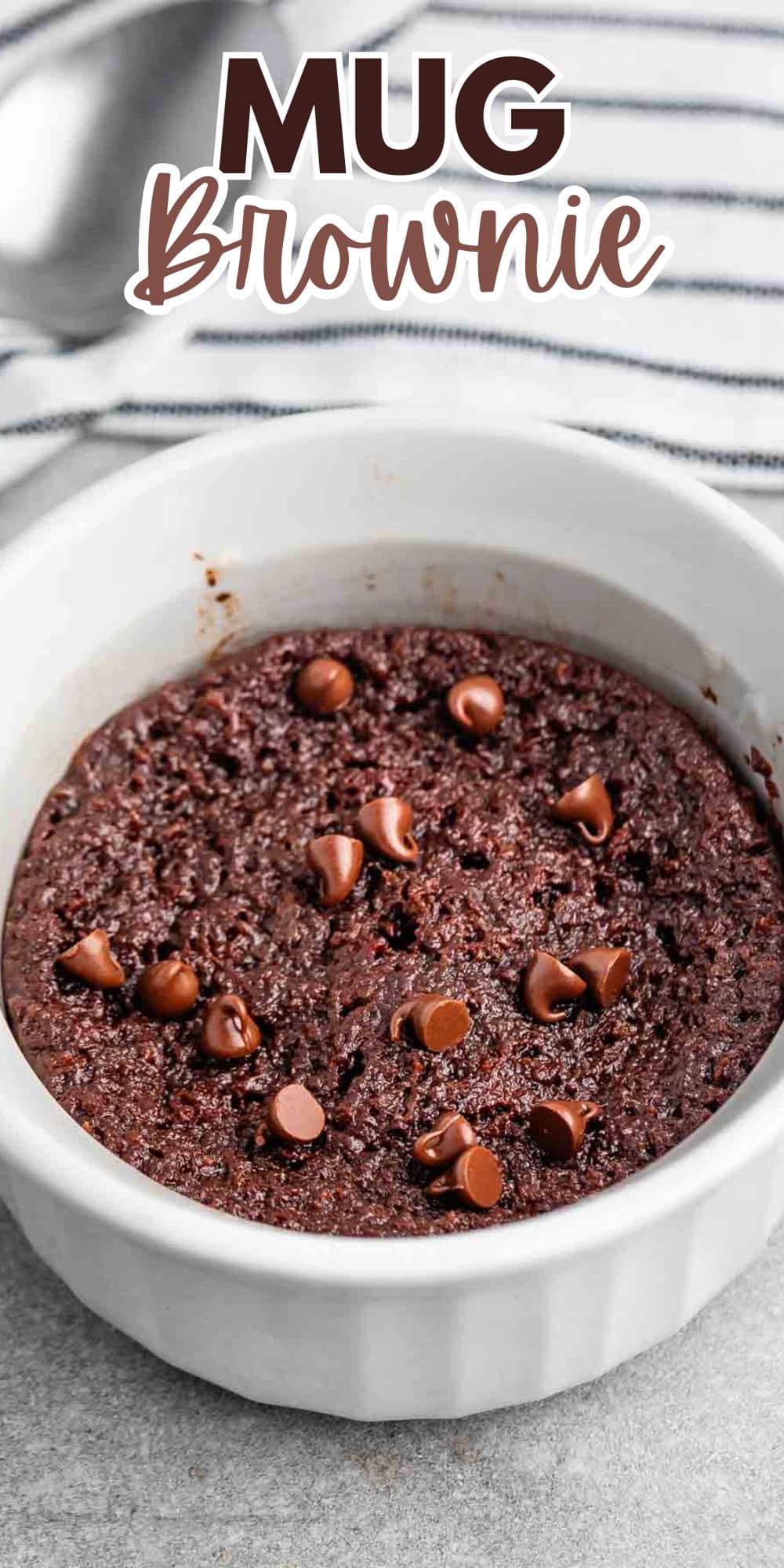 A chocolate mug brownie topped with chocolate chips in a white ramekin. A striped cloth and metal spoon are in the background.