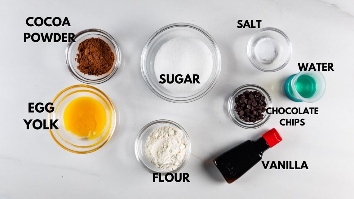 Image of baking ingredients on a white surface: small bowls of cocoa powder, sugar, salt, egg yolk, and flour, along with chocolate chips, a glass of water, and a bottle of vanilla extract, all labeled with text.