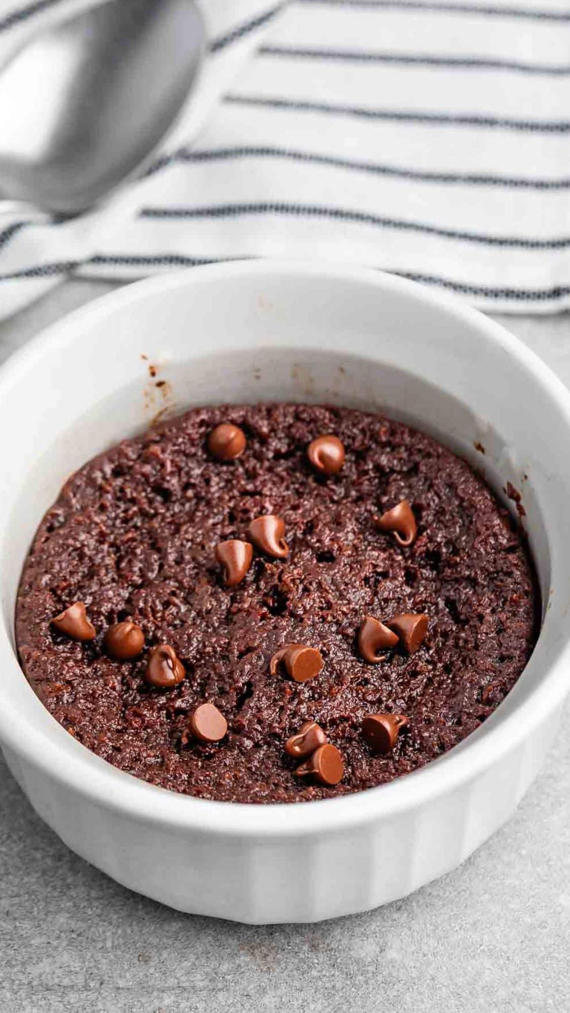 A small white ramekin filled with a freshly baked chocolate mug cake, topped with chocolate chips. A striped kitchen towel and a spoon are blurred in the background.