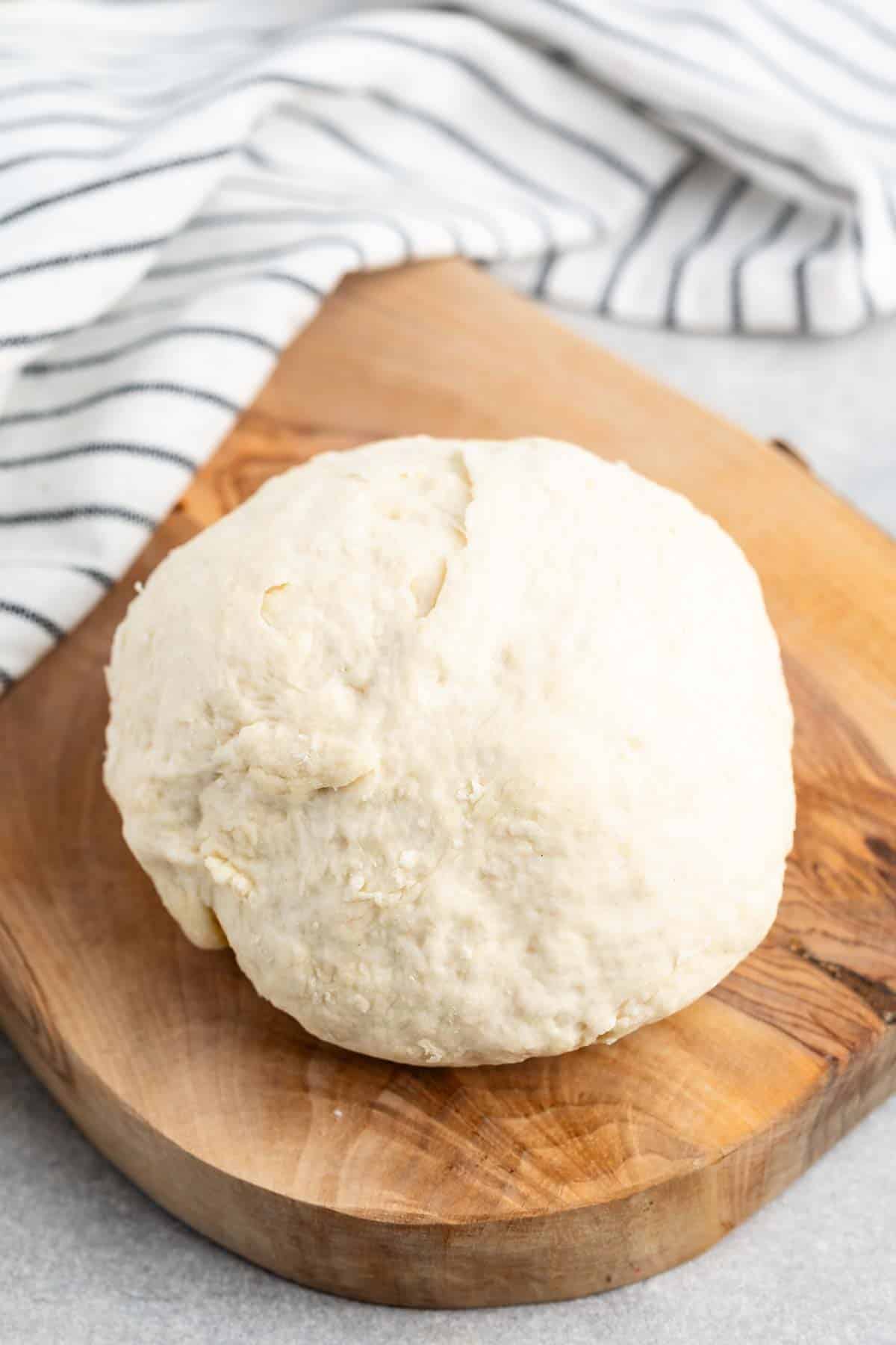 A ball of raw dough sits on a wooden cutting board. The dough appears freshly kneaded, with a smooth texture. In the background, a striped kitchen towel is partially visible on a gray surface.