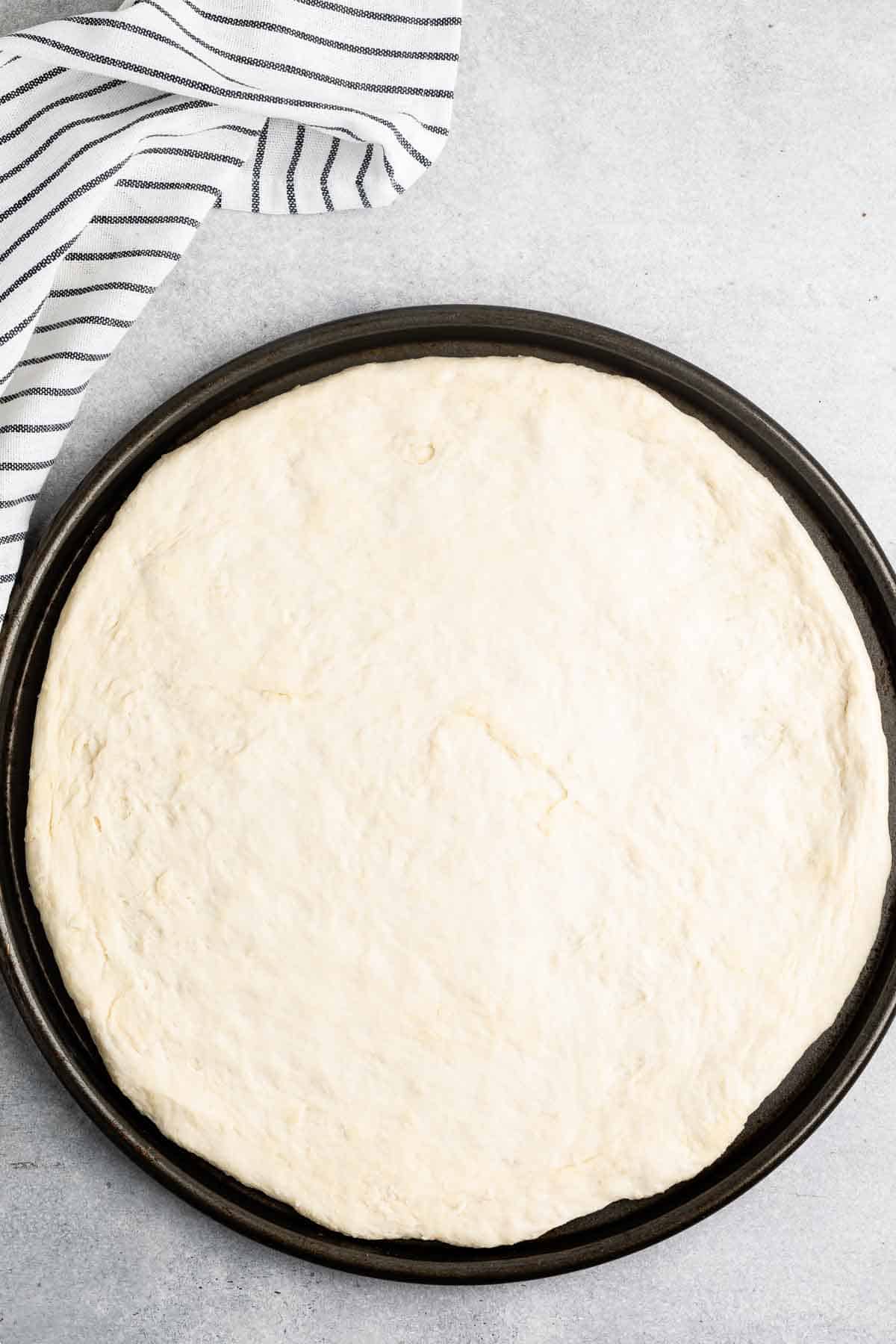 A round pizza dough neatly spread on a black baking tray, placed on a light gray countertop. A white kitchen towel with black stripes is partially visible in the top left corner of the image.