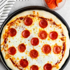 A freshly baked pepperoni pizza on a round black tray, topped with melted cheese and pepperoni slices. A small clear bowl with extra pepperoni is beside the pizza. A striped cloth is partially visible in the top left corner.