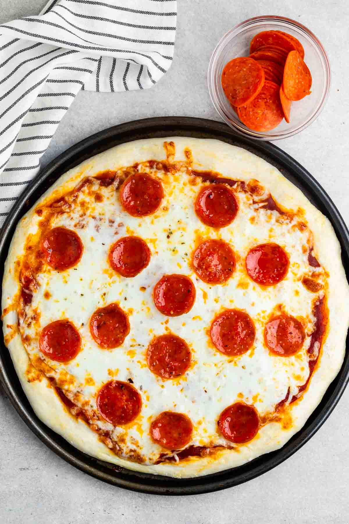 A freshly baked pepperoni pizza on a round black tray, topped with melted cheese and pepperoni slices. A small clear bowl with extra pepperoni is beside the pizza. A striped cloth is partially visible in the top left corner.