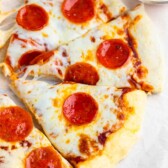 Close-up of a pepperoni pizza with three slices arranged on parchment paper. Melted cheese covers the slices, each topped with several pepperoni slices. A small bowl with extra pepperoni is visible in the background.