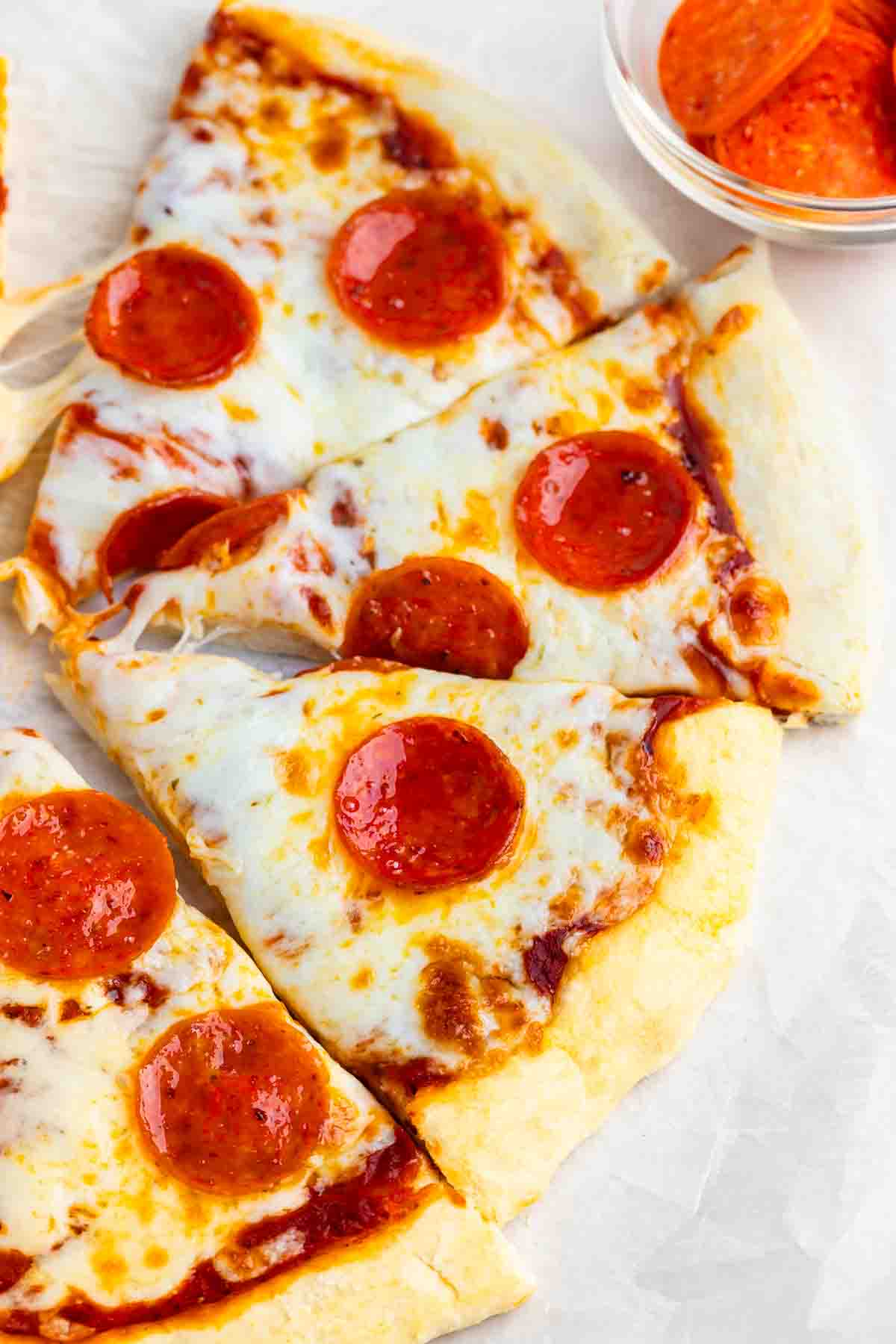 Close-up of a pepperoni pizza with three slices arranged on parchment paper. Melted cheese covers the slices, each topped with several pepperoni slices. A small bowl with extra pepperoni is visible in the background.