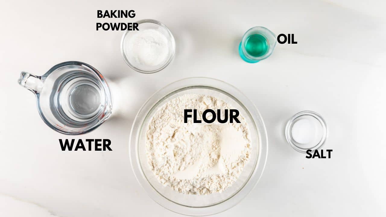 Top-down view of baking ingredients on a white surface, including a glass bowl of flour, small dishes of baking powder and salt, a measuring cup of water, and a small container of oil. Each item is labeled with its name.