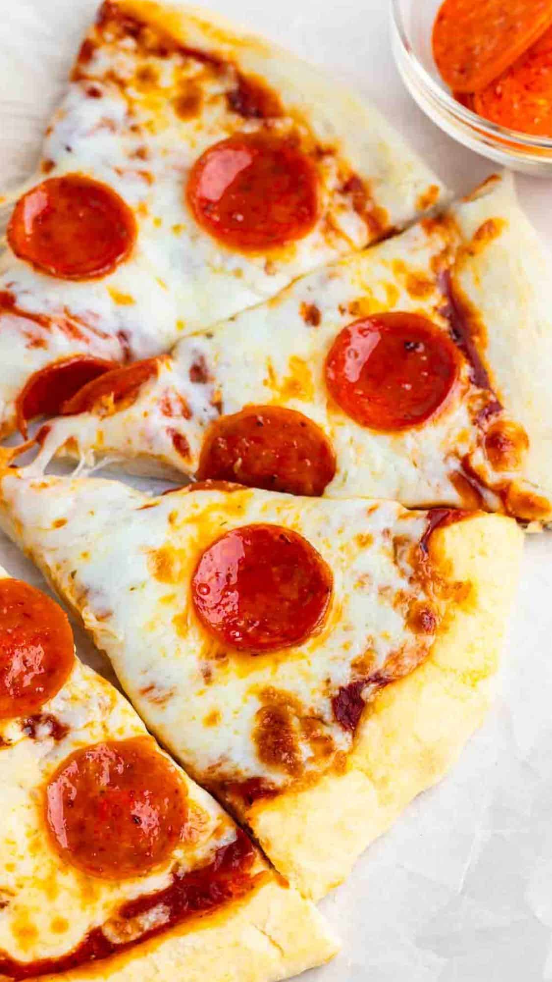 Slices of pepperoni pizza with melted cheese and tomato sauce on a light-colored surface. A small bowl with additional pepperoni slices is visible in the background.