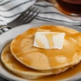 A stack of pancakes topped with a pat of butter and drizzled with syrup on a white plate. A fork and a glass container of syrup are in the background, placed on a striped cloth.