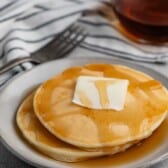 A stack of pancakes topped with a pat of butter and drizzled with syrup on a white plate. A glass syrup container and a striped napkin with a fork are in the background.