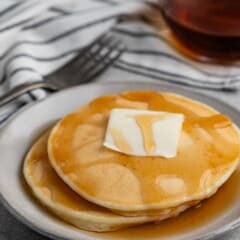 A stack of pancakes topped with a pat of butter and drizzled with syrup on a white plate. A glass syrup container and a striped napkin with a fork are in the background.