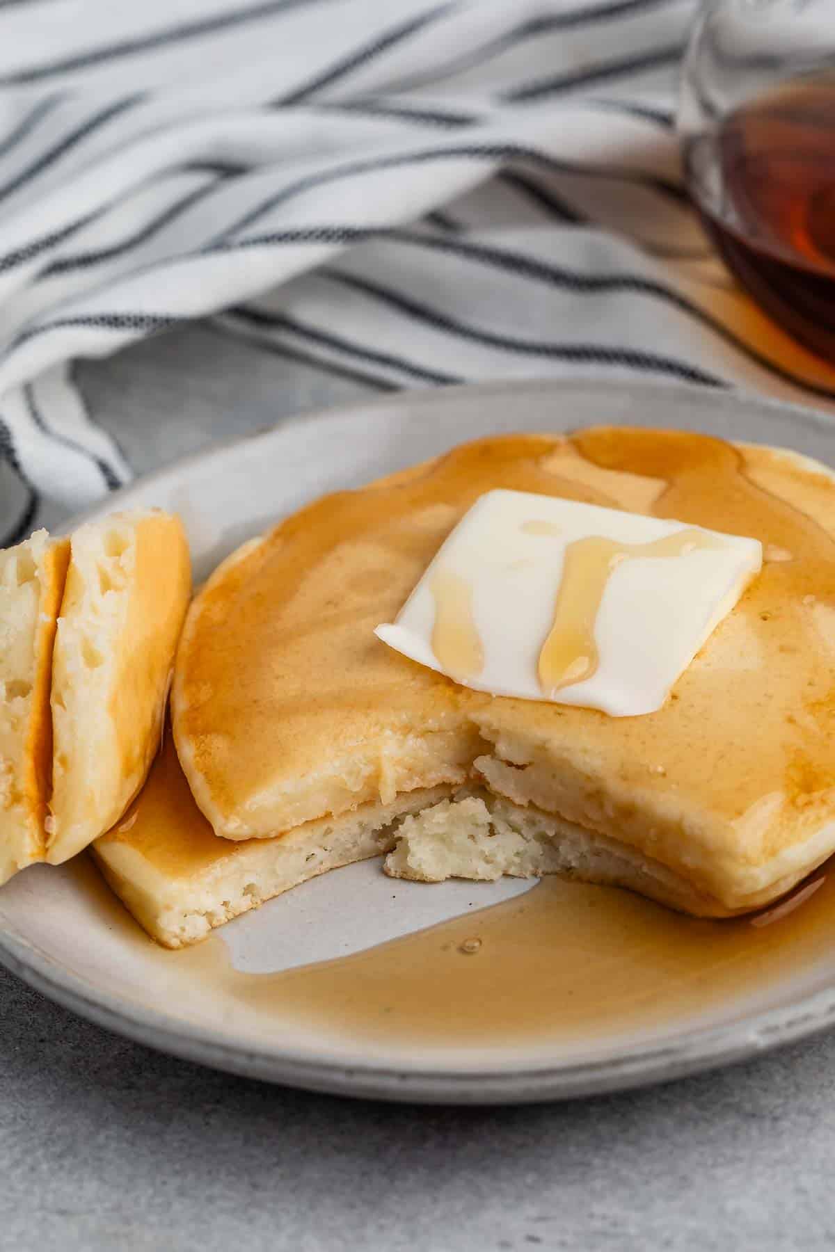 A stack of pancakes on a plate with a slice of butter melting on top, drizzled with syrup. A piece of pancake is cut out, and a glass container of syrup is in the background. A striped cloth adds texture to the scene.