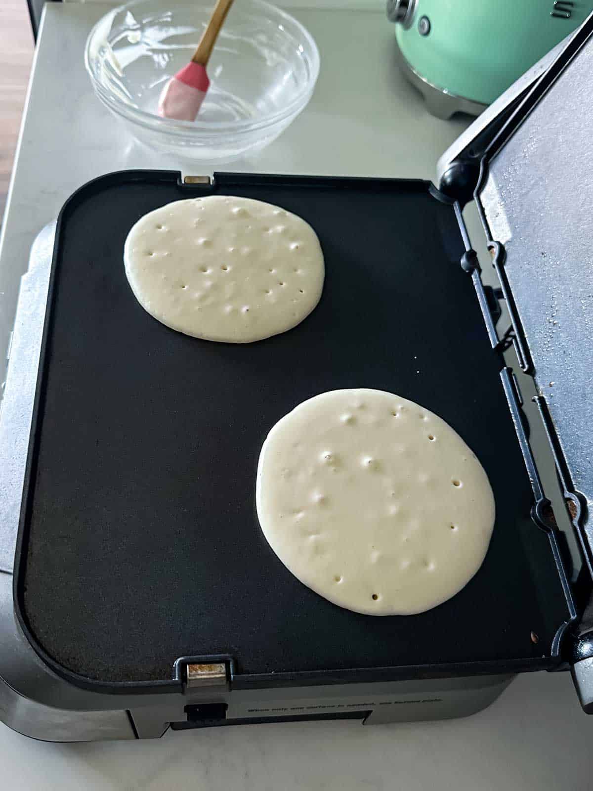 Two pancakes cooking on a black griddle, each with small bubbles on the surface. A clear bowl with a red-handled brush is in the background on a light-colored countertop.