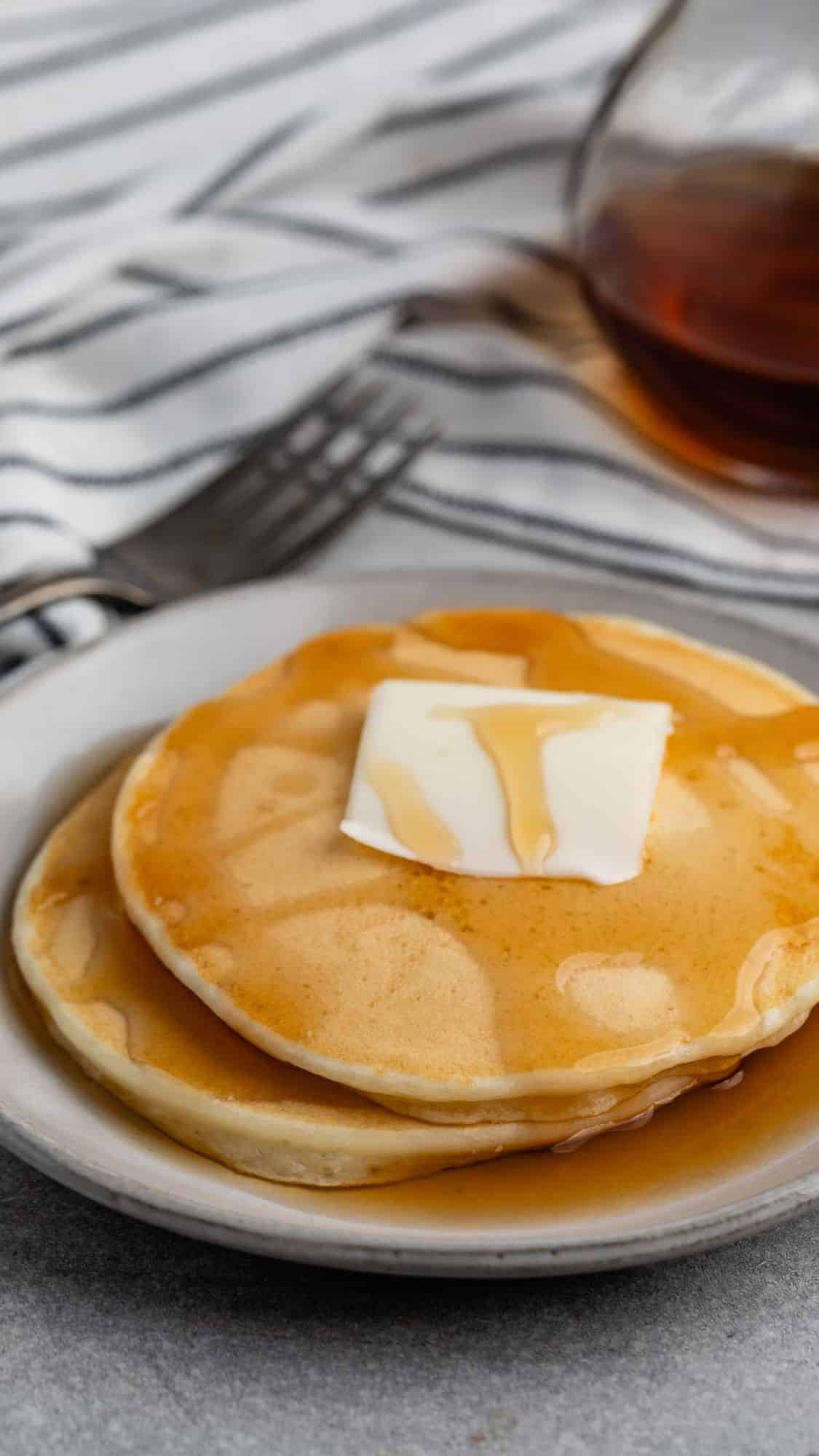 A stack of pancakes topped with a pat of butter and drizzled with syrup on a white plate. A glass syrup pitcher and a fork are in the background on a striped cloth.