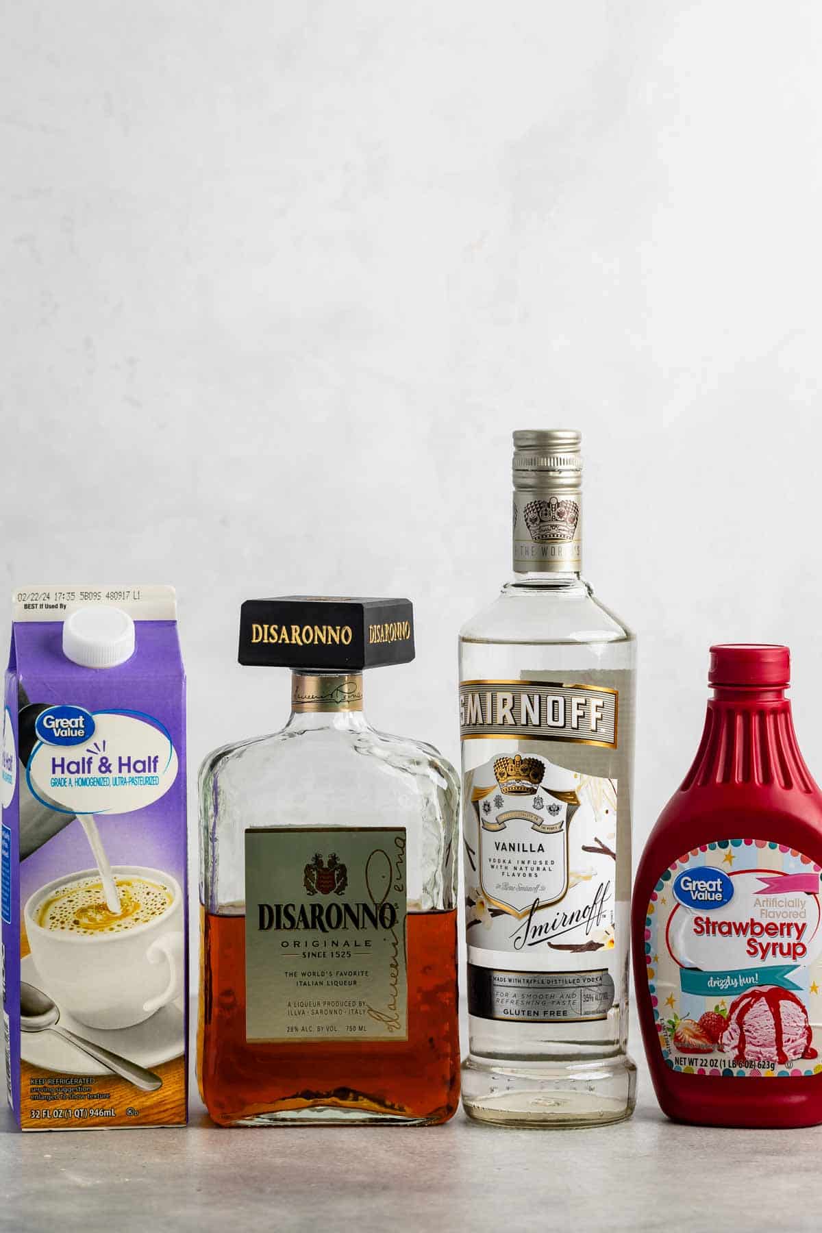 A selection of ingredients on a countertop: a carton of half and half, a bottle of Disaronno liqueur, a bottle of Smirnoff Vanilla vodka, and a bottle of strawberry syrup.