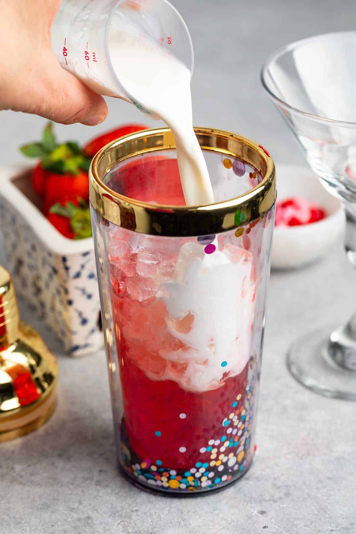 A hand pours milk into a tall glass filled with ice and pink liquid. The glass has colorful dots and a gold rim. Strawberries and another glass are in the background on a gray surface.