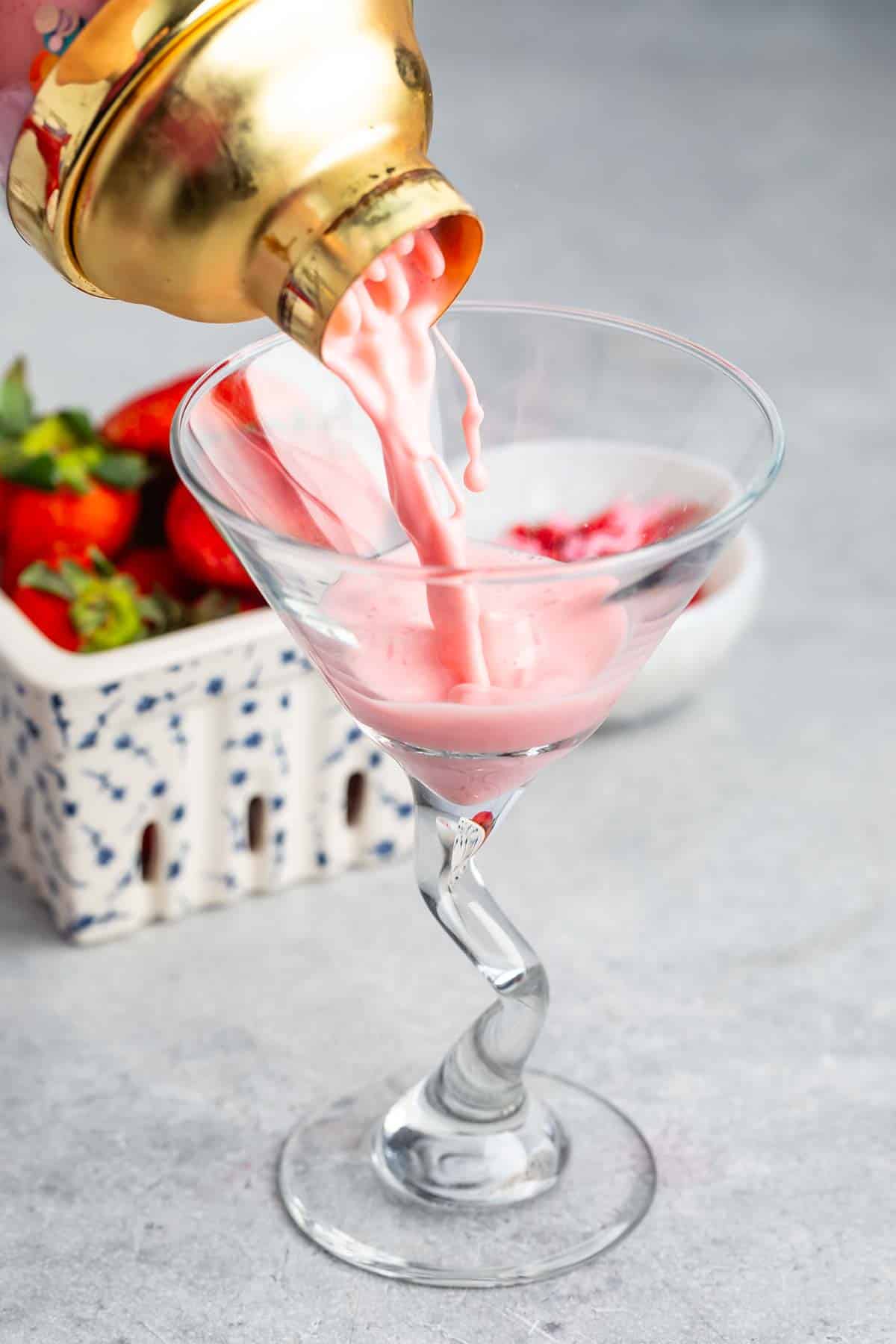 A pink cocktail is being poured from a golden shaker into a uniquely twisted martini glass. In the background, there is a white container with fresh strawberries. The setting is on a gray surface.