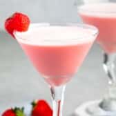 Two pink cocktails in martini glasses, each topped with a strawberry on the rim. More strawberries are scattered on the gray surface. The background is blurred, highlighting the drinks.