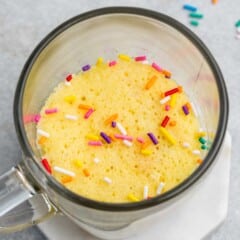 A glass mug on a hexagonal white coaster contains a fluffy yellow cake topped with colorful sprinkles. Scattered sprinkles are visible on the gray surface around the mug.