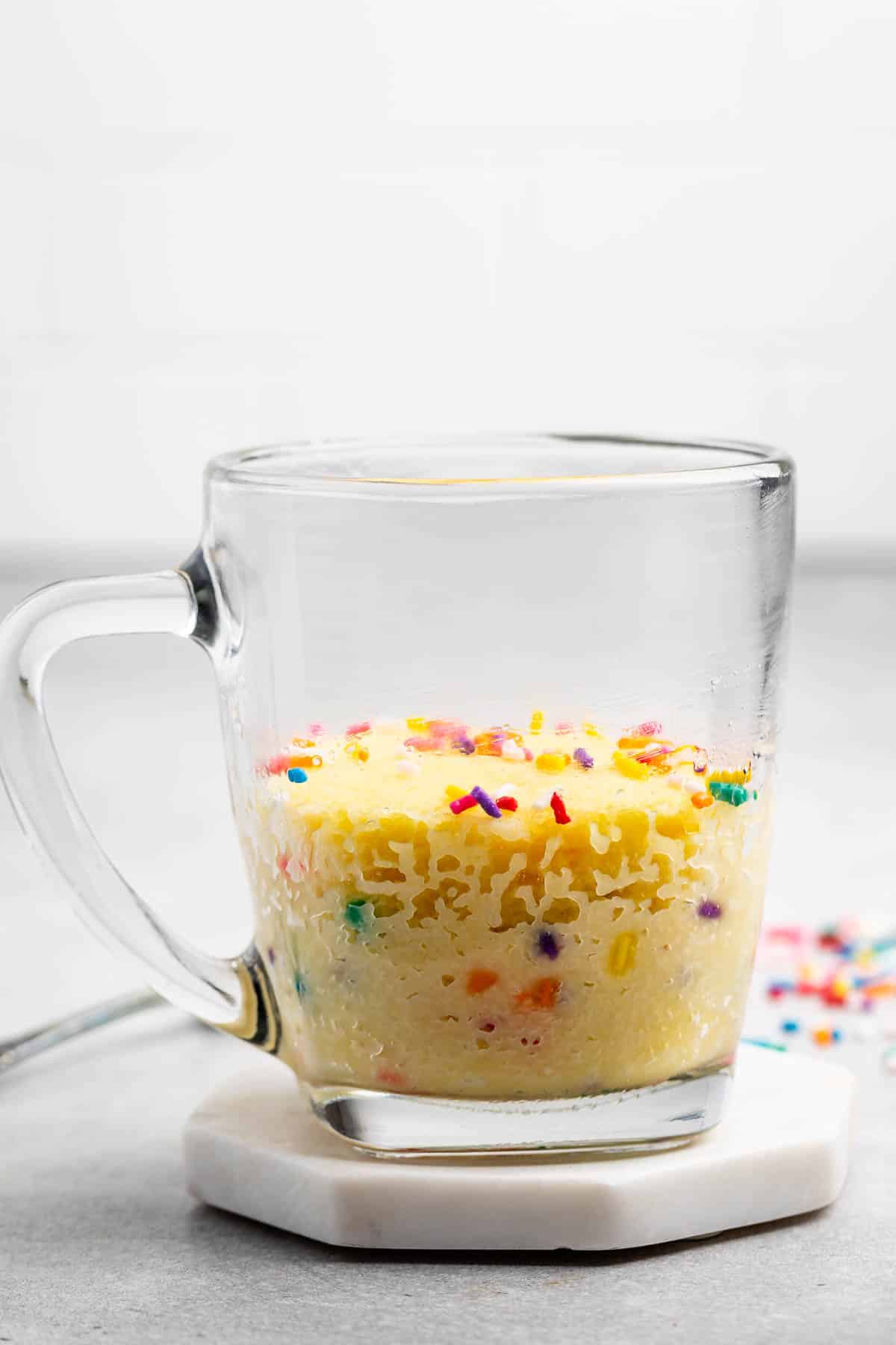 A clear glass mug on a white coaster contains a mug cake with colorful sprinkles scattered on top. The mug is placed on a gray surface with a few sprinkles around it, and a blurred white background is visible.