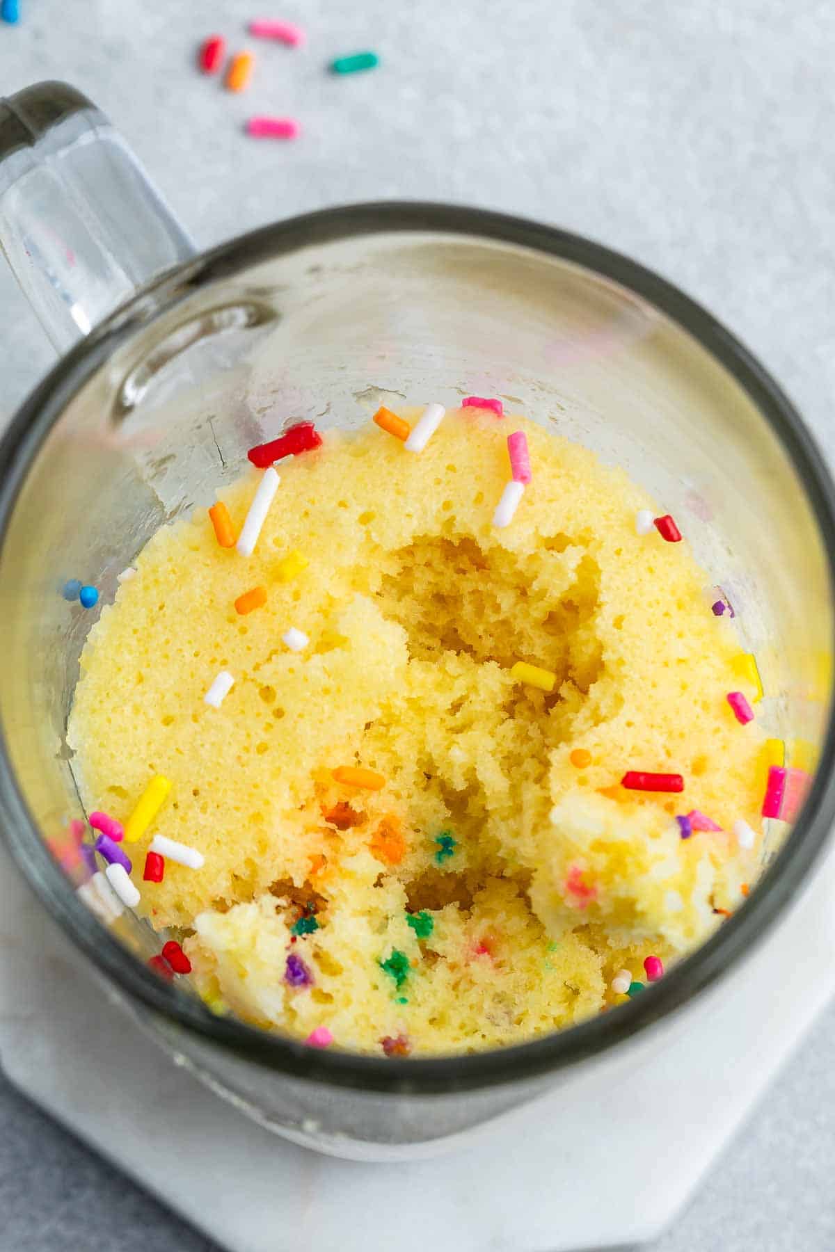 A close-up of a vanilla mug cake in a glass mug, topped with colorful sprinkles. One spoonful is missing, revealing the moist texture of the cake. The mug rests on a light-colored surface with scattered sprinkles nearby.