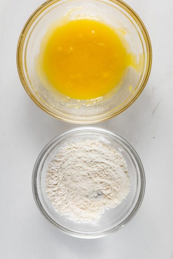 Two glass bowls are on a white surface. The left bowl contains a beaten yellow egg mixture, and the right bowl is filled with white flour.