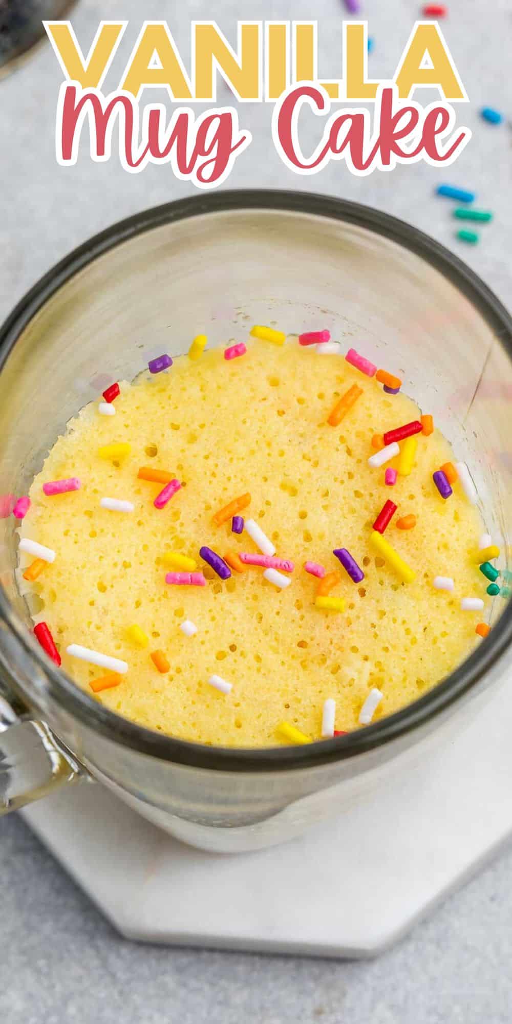 A mug filled with a fluffy vanilla cake topped with colorful sprinkles. The mug is placed on a marble surface. The text Vanilla Mug Cake appears at the top of the image.