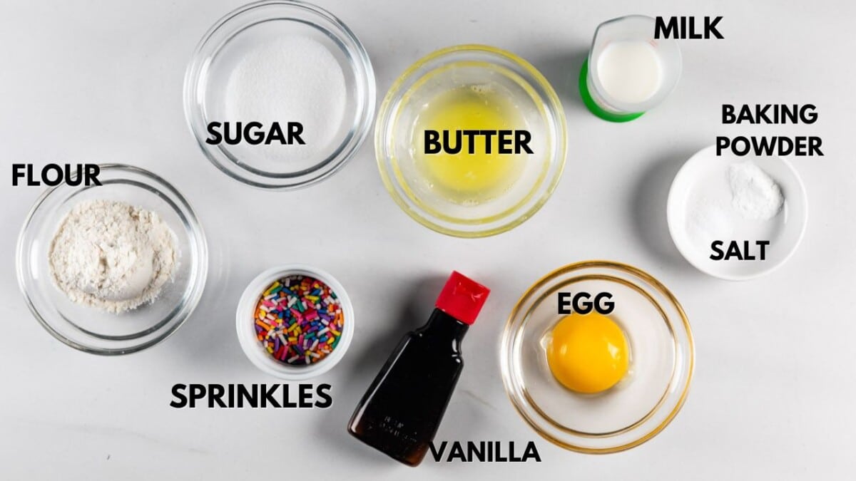 A top-view image of ingredients in separate bowls on a white surface. They include flour, sugar, melted butter, milk, baking powder, salt, an egg yolk, colorful sprinkles, and a bottle of vanilla extract. Each ingredient is labeled.