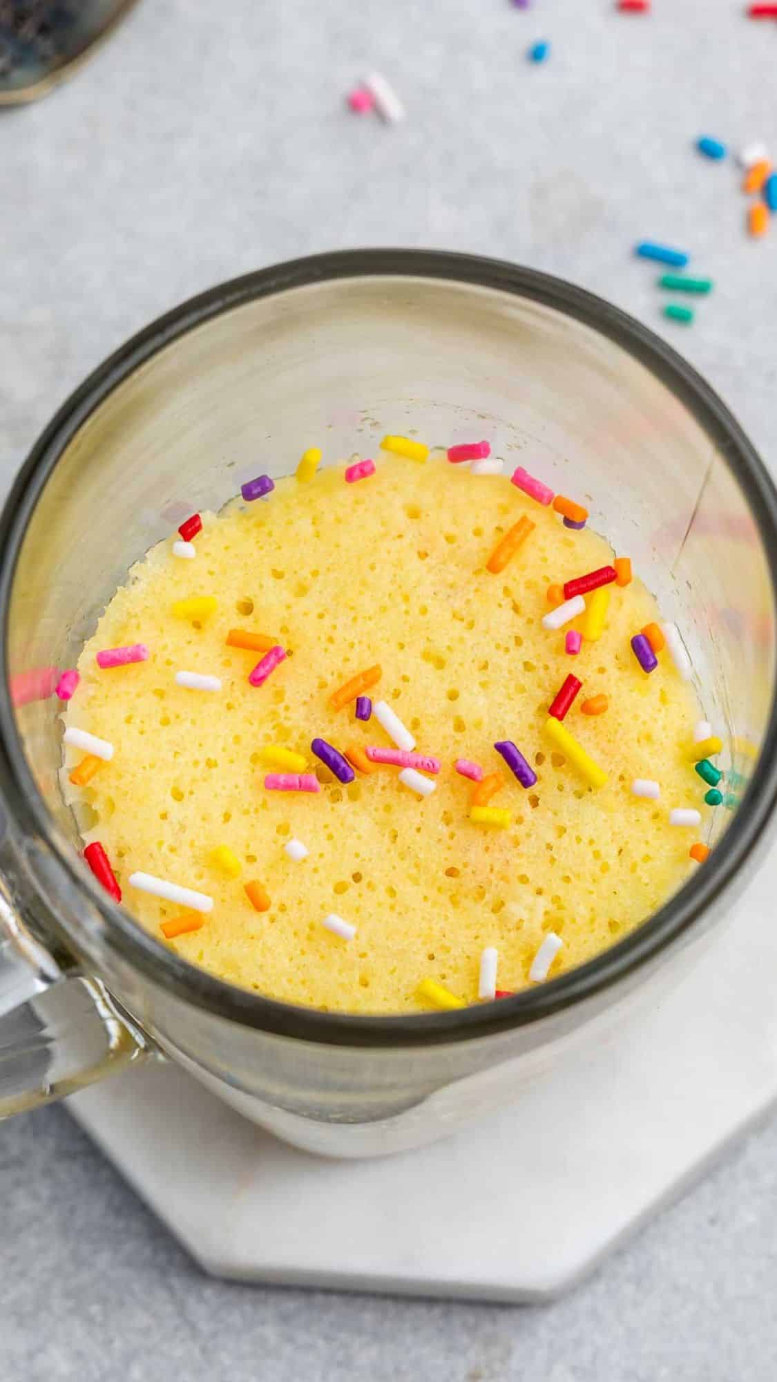 A mug cake topped with colorful sprinkles sits in a clear glass mug. The background features a gray countertop with more scattered sprinkles, and the mug is placed on a white, hexagonal coaster.