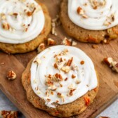 Carrot cake cookies with white frosting swirl and chopped nuts are displayed on a wooden board. A green bowl with more nuts is in the background. The cookies have visible carrot pieces and a soft, rustic appearance.