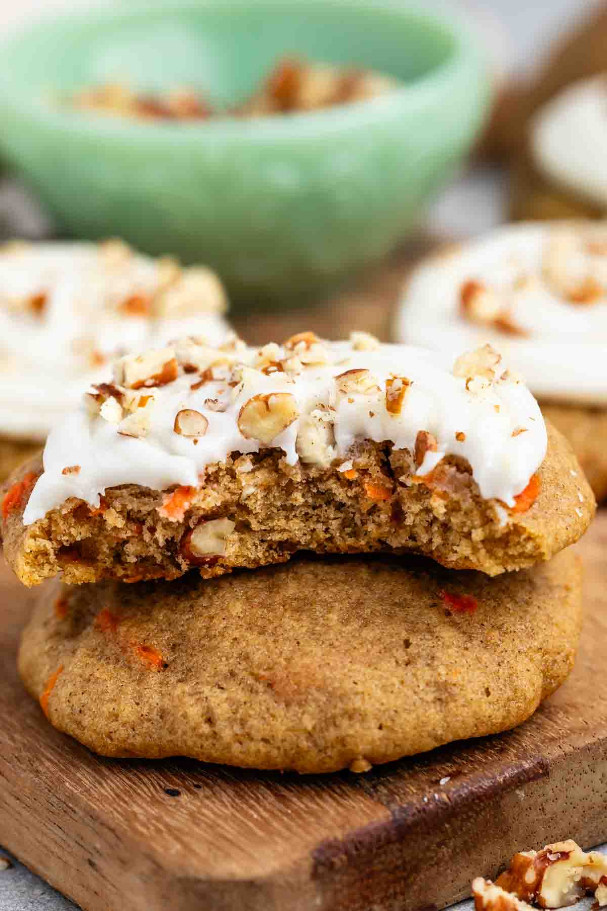 Carrot cake cookies topped with cream cheese frosting and chopped pecans sit on a wooden board. One cookie is broken in half, showing its moist interior. A green bowl is blurred in the background.