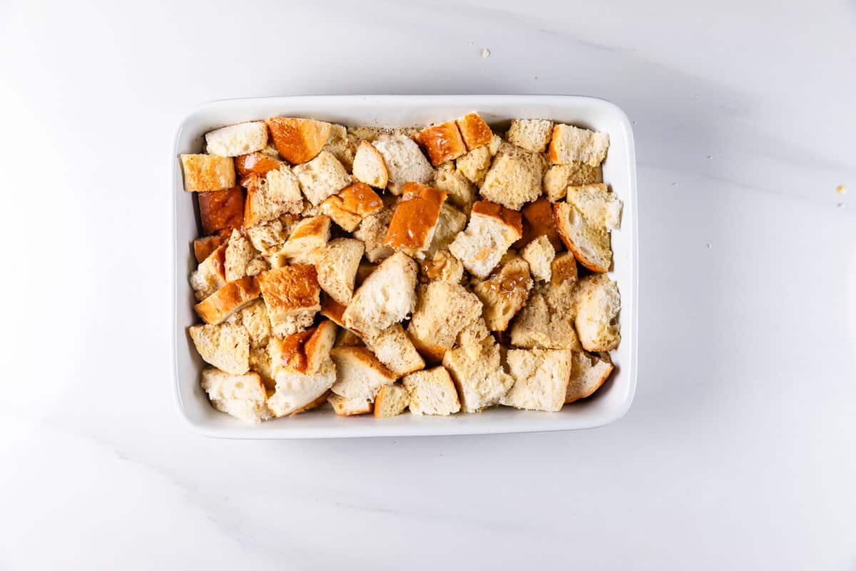 A white baking dish filled with cubed pieces of bread, scattered randomly. The bread appears slightly toasted, suggesting preparation for a dish like stuffing or bread pudding. The background is a smooth, white surface.