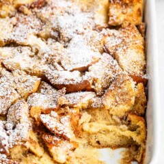 A baked bread pudding in a rectangular white dish, topped with a dusting of powdered sugar. Some pieces are golden-brown and slightly crispy. A section is missing, revealing the soft, custardy interior.