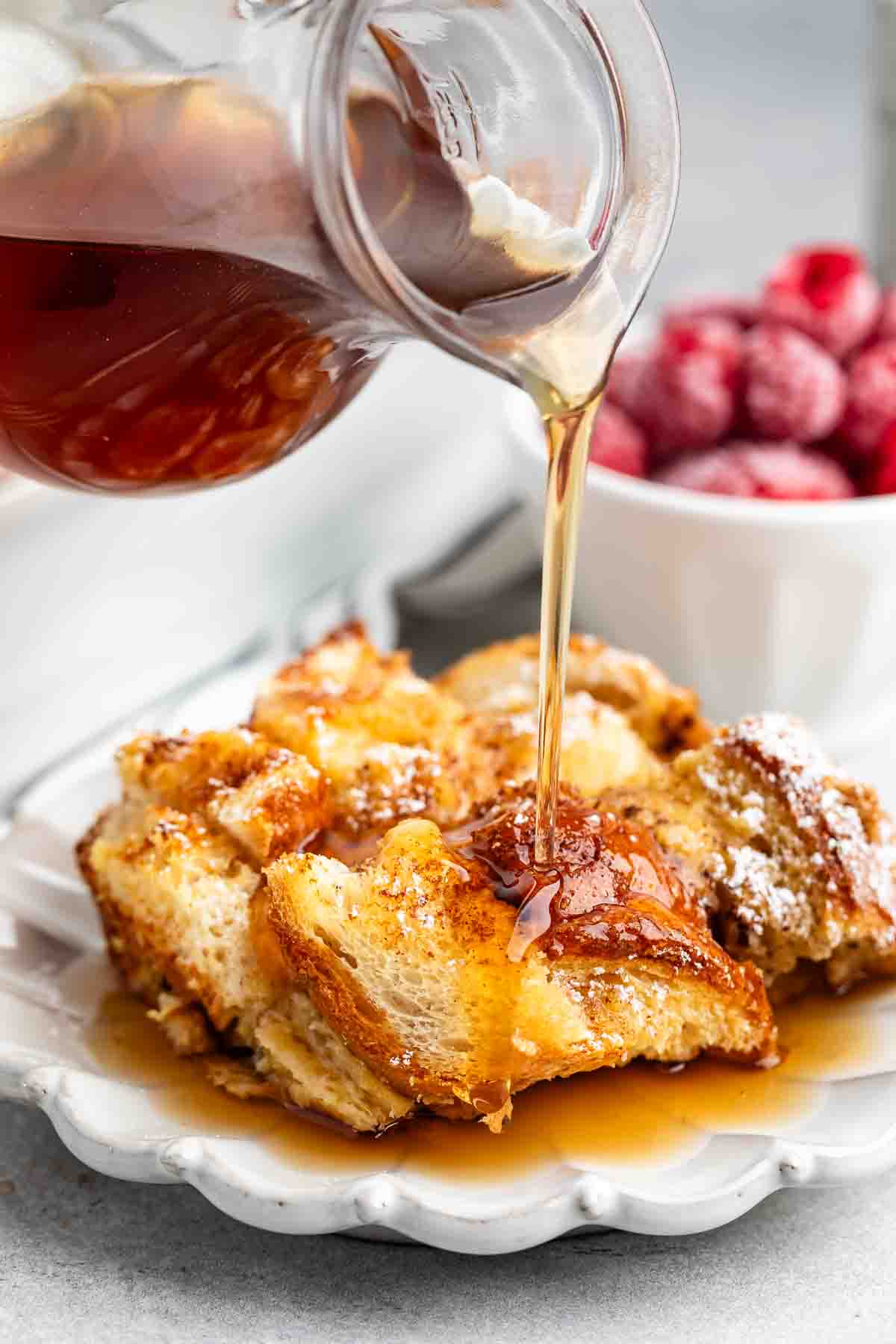 Syrup is being poured from a glass pitcher over a plate of French toast, which is sprinkled with powdered sugar. In the background, a bowl of fresh raspberries is visible.