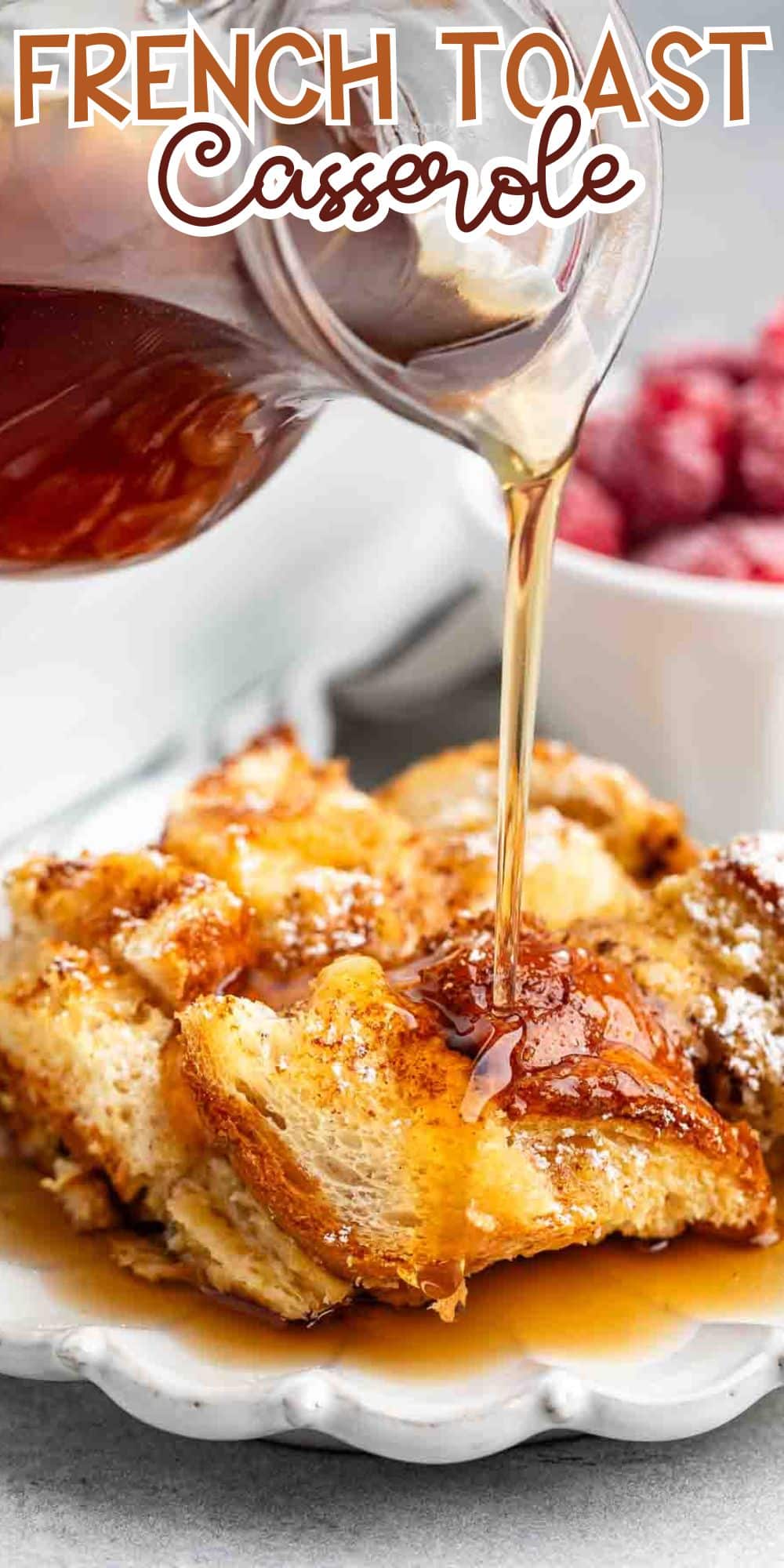 A close-up of French toast casserole on a plate, with syrup being poured over it. In the background, there is a bowl of strawberries. The words French Toast Casserole are written at the top.