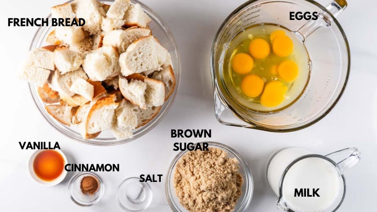 Ingredients for a recipe are arranged on a table. Clockwise from top left: a bowl of French bread pieces, a pitcher with eggs, a small bowl of milk, a bowl of brown sugar, and small bowls containing salt, cinnamon, and vanilla.