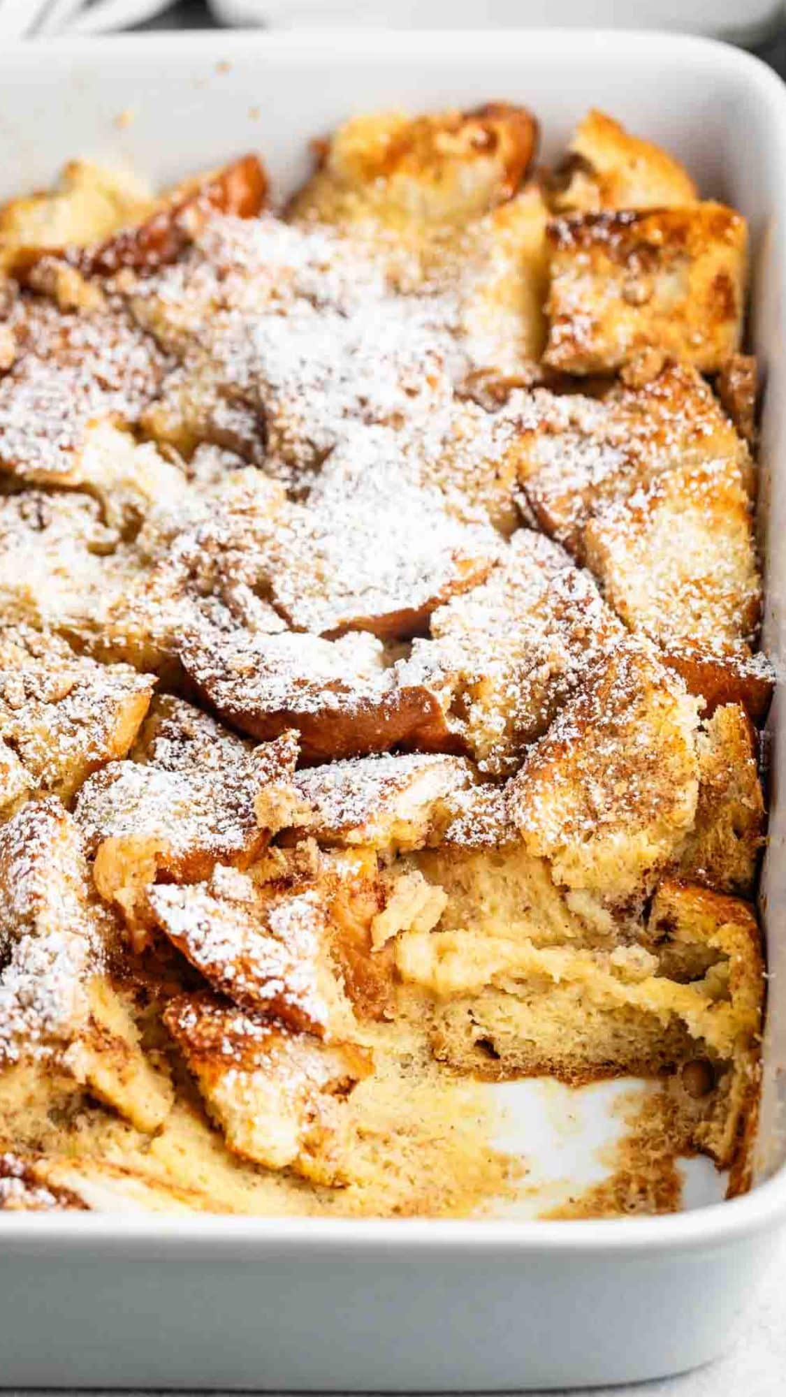 A baking dish filled with golden brown French toast casserole, dusted with powdered sugar. Some pieces are missing, revealing a creamy interior.