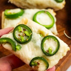 A hand holding a slice of bread topped with melted cheese and sliced jalapeños. Additional slices are visible in the background on a wooden surface.