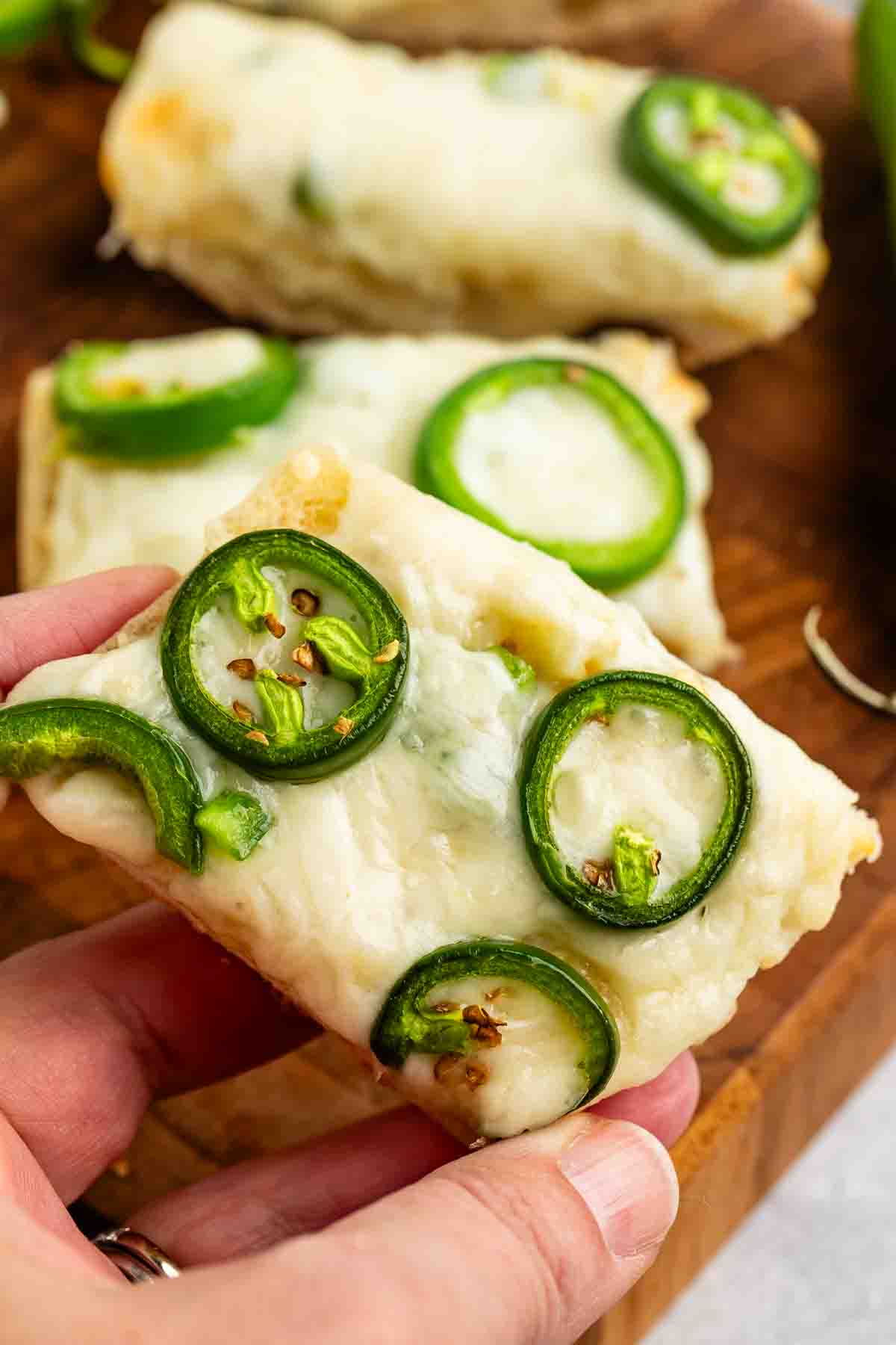 A hand holding a slice of bread topped with melted cheese and sliced jalapeños. Additional slices are visible in the background on a wooden surface.