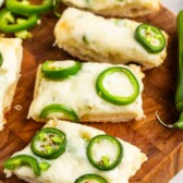 Rectangular slices of bread topped with melted cheese and sliced jalapeños are arranged on a wooden cutting board. A few loose jalapeño slices are scattered nearby.