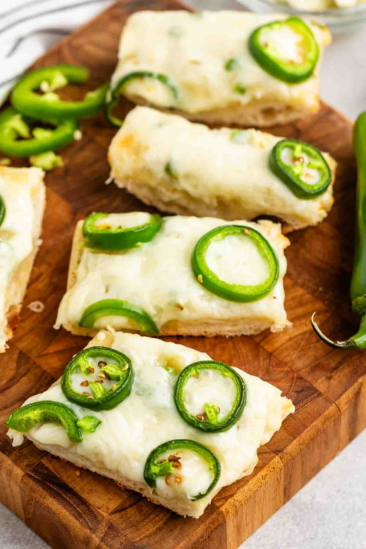 Rectangular slices of bread topped with melted cheese and sliced jalapeños are arranged on a wooden cutting board. A few loose jalapeño slices are scattered nearby.