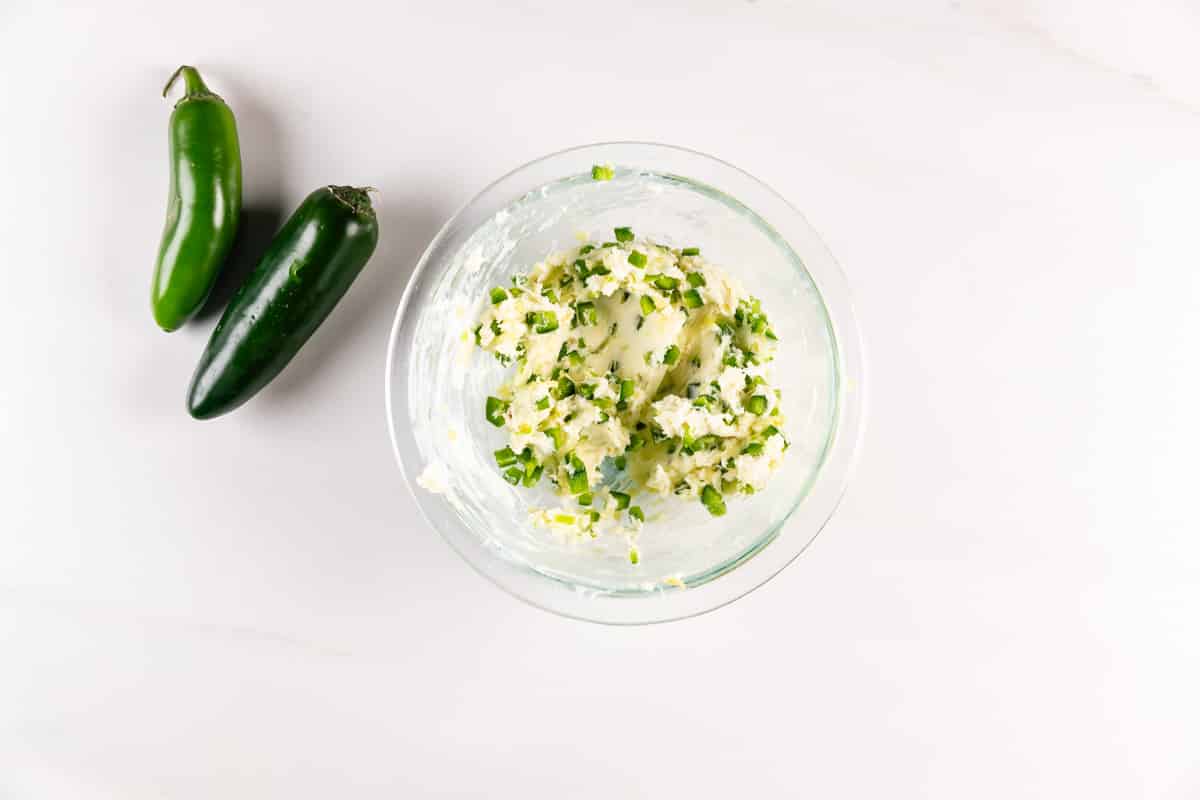 Bowl of jalapeño butter with chopped green herbs on a white surface. Two whole jalapeños are placed next to the bowl.