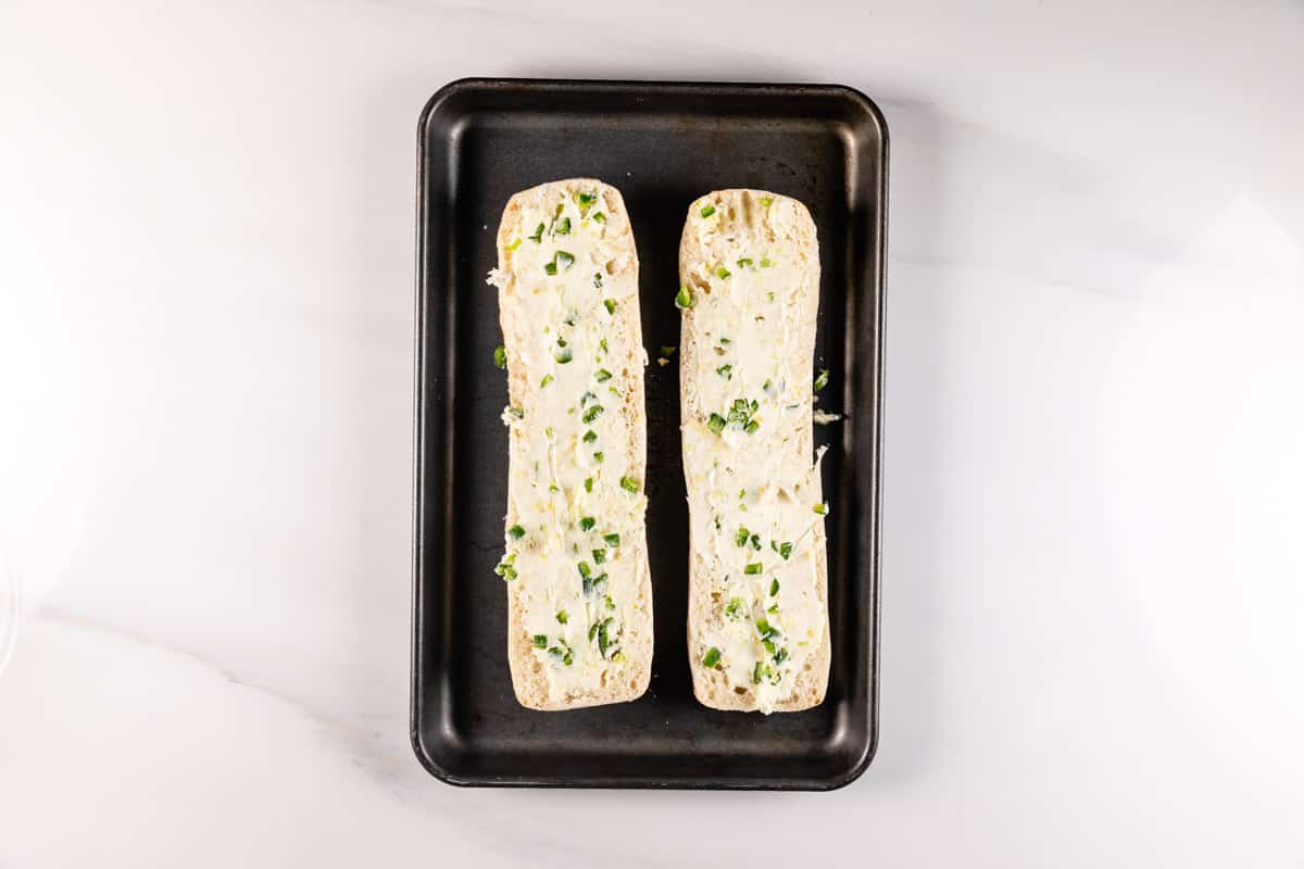 A baking tray holding two halves of a baguette topped with garlic butter and chopped green herbs, ready to be baked. The tray is placed on a white marble surface.