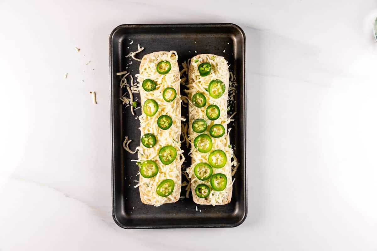 Two uncooked baguettes topped with shredded cheese and sliced jalapeños on a black baking tray. The tray is placed on a light marble surface. Some cheese shreds are scattered around the tray.