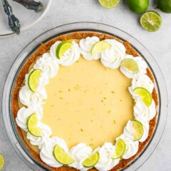 A Key lime pie garnished with whipped cream and lime slices sits on a gray surface. Beside the pie are whole and halved limes. Two decorative spoons rest on a stack of white plates in the background.